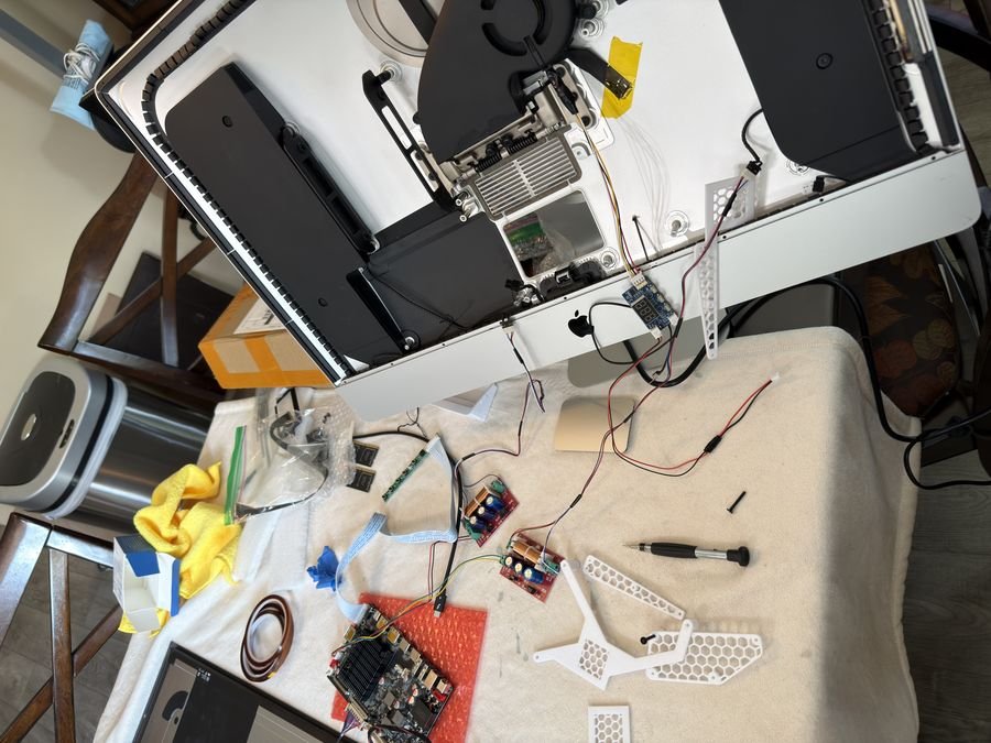 iMac chassis tilted up on a work table with the driver board and 3D printed brackets being fitted, crossover boards and tools spread out below